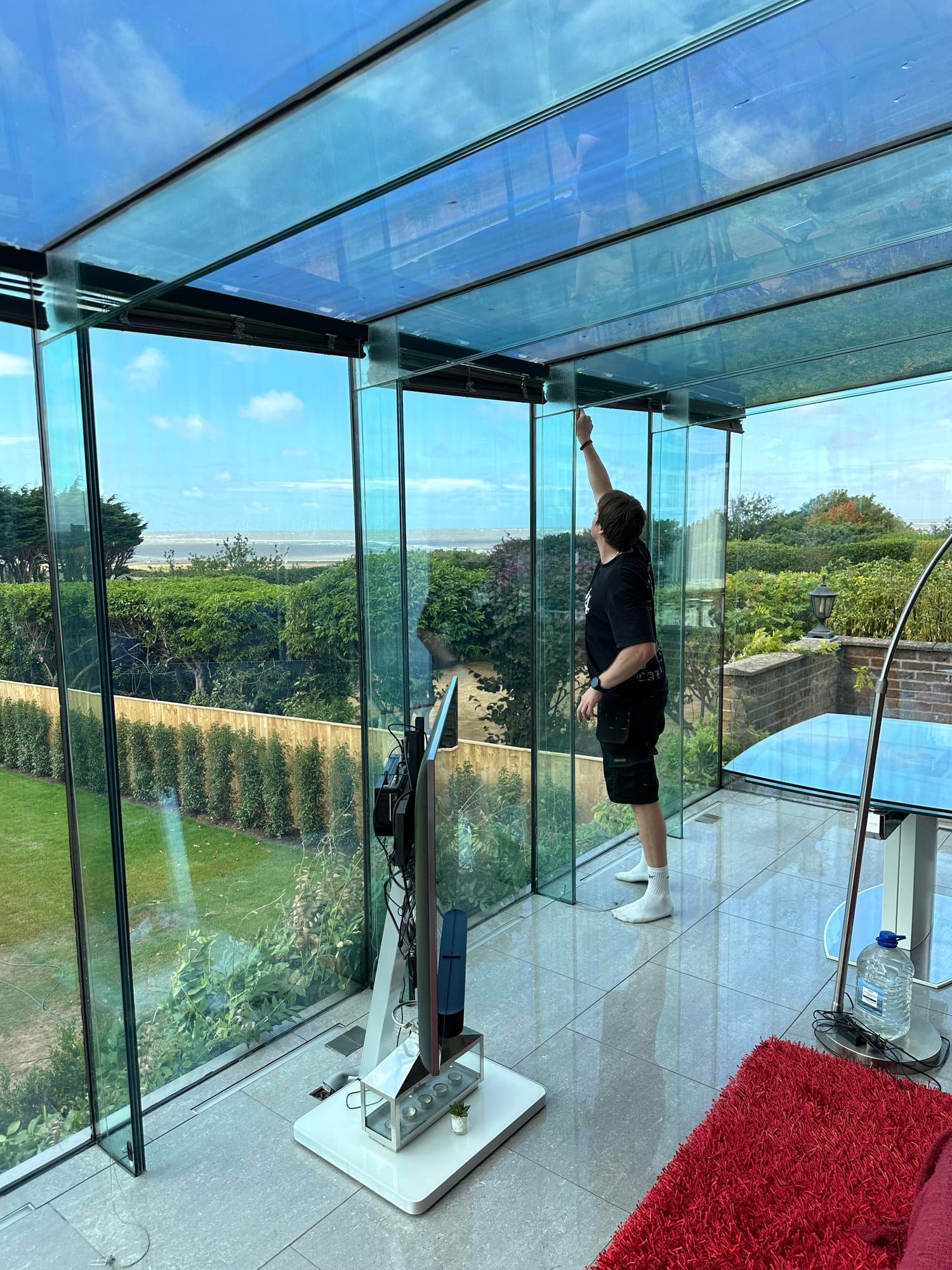An Eclipse Shield fitter applying window film to a frameless glass room with a garden and sea view behind