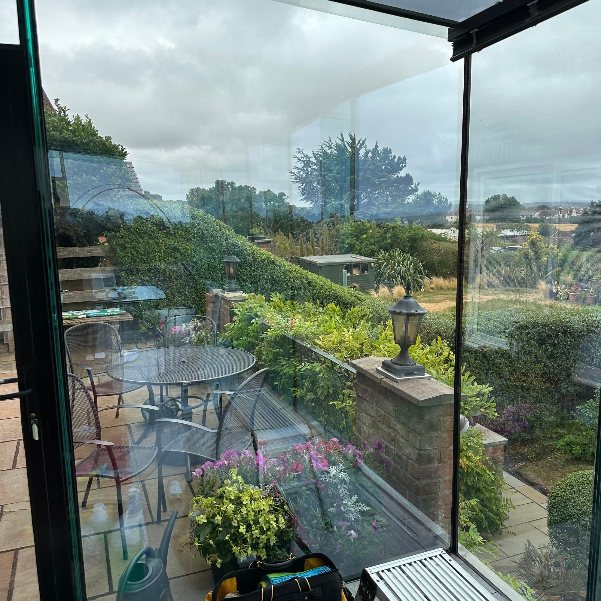 View out through the floor-to-ceiling glass of a garden room onto a terrace with table and chairs